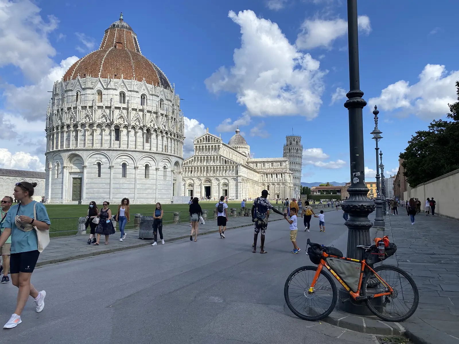 The loaded bike parked beside a lamp post in the Piazza dei Miracoli, with the Baptistery, Duomo and Leaning Tower of Pisa behind crowds of visitors.