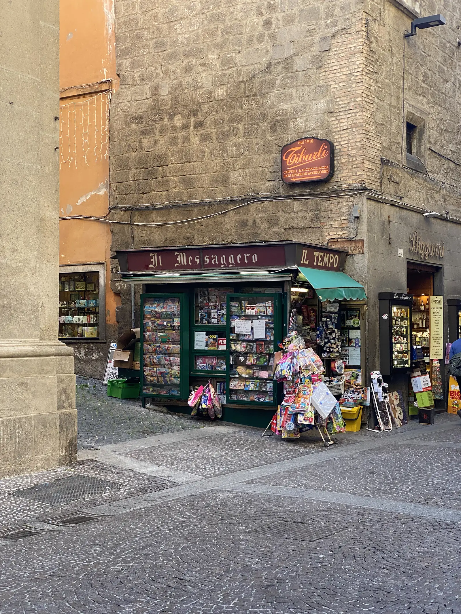 A newsstand kiosk labelled "Il Messaggero" and "Il Tempo" packed with magazines and toys on a cobbled street corner, a "Tiburli" hat and accessories shop sign on the building above.