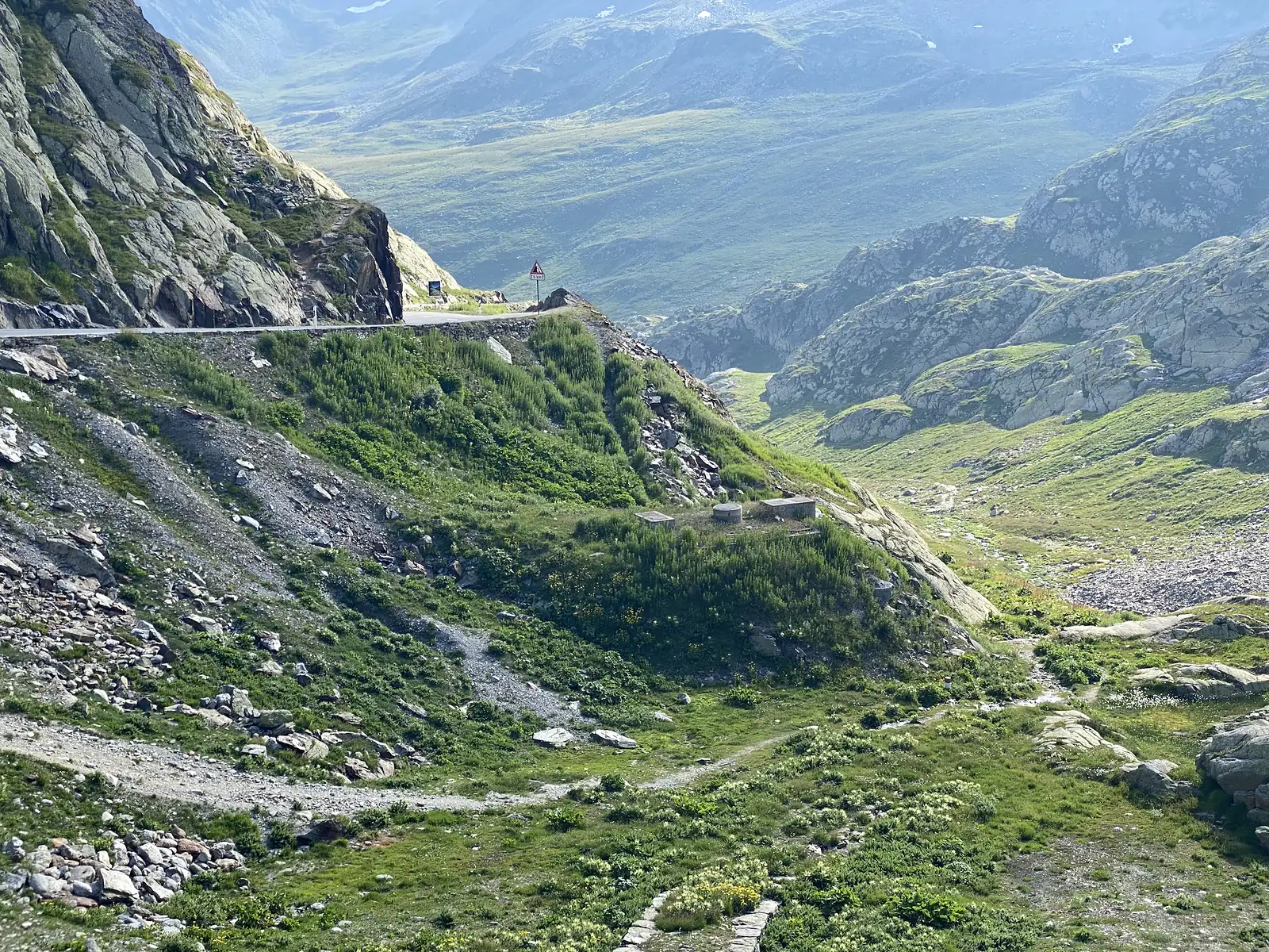 A stony path descends a mountainside past a red-and-white warning sign, with a green valley opening out below.