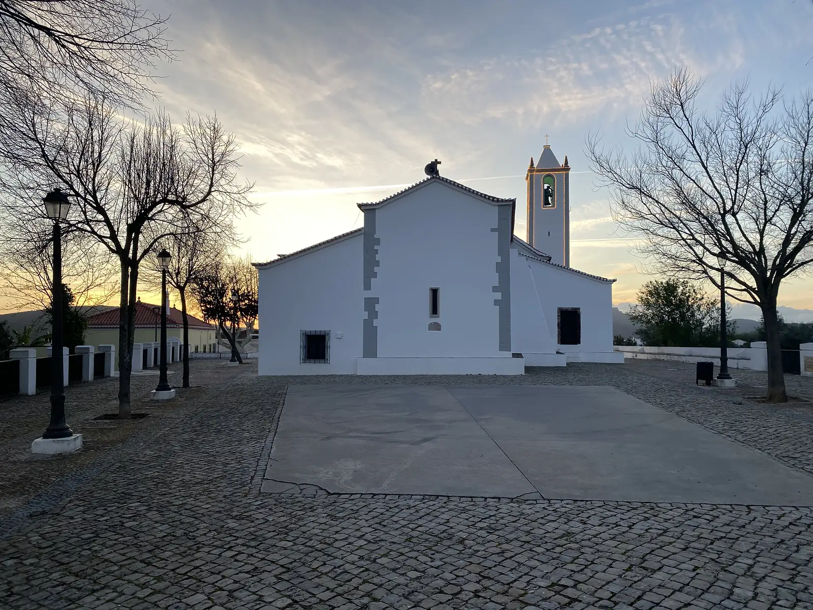 Whitewashed village church with a stepped bell tower and a single bell, seen across an empty cobbled square with lamp posts and leafless trees, the sky flushed with sunset behind it.