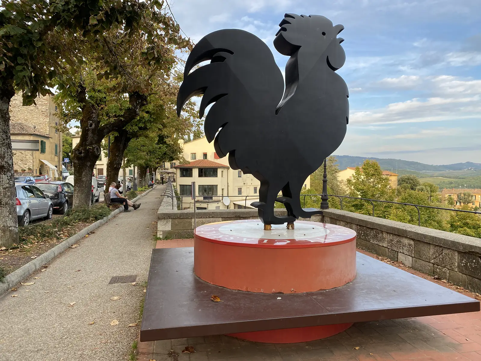 A large black rooster statue, the Chianti Classico emblem, mounted on a red plinth on a paved terrace overlooking distant hills.