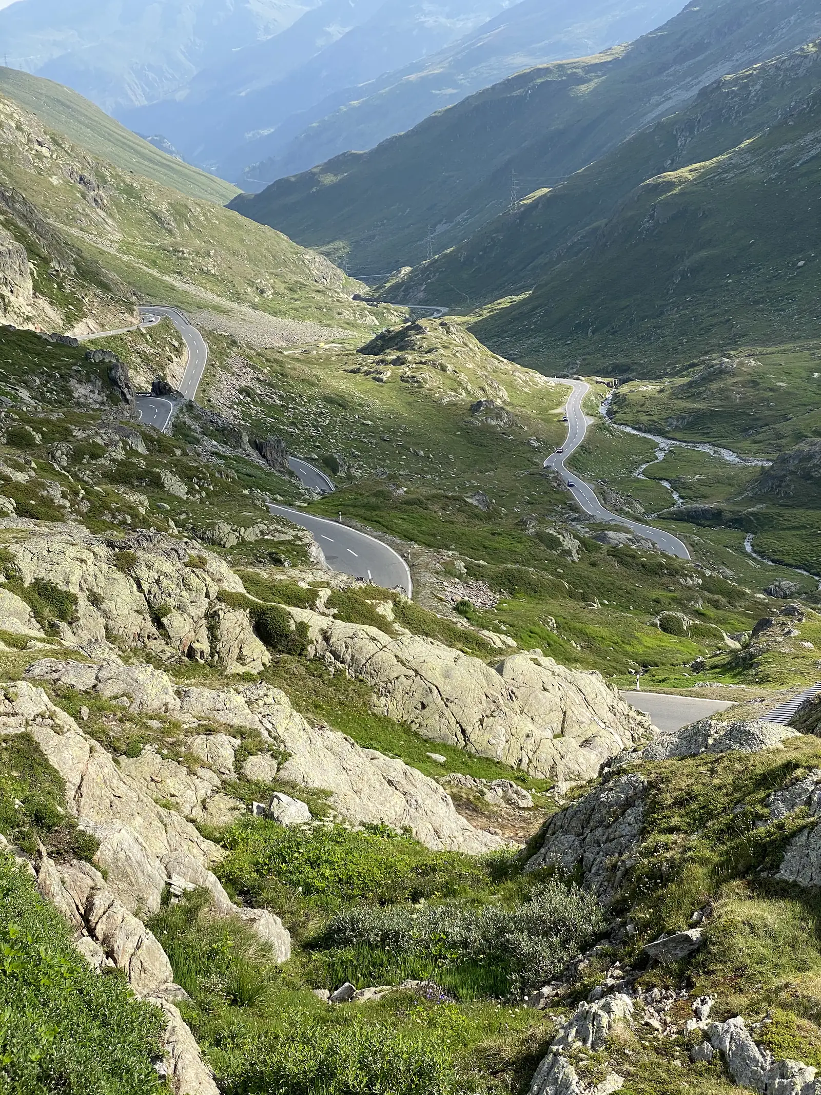 A mountain road switches back down a valley in a series of hairpin bends, flanked by rocky grassland.
