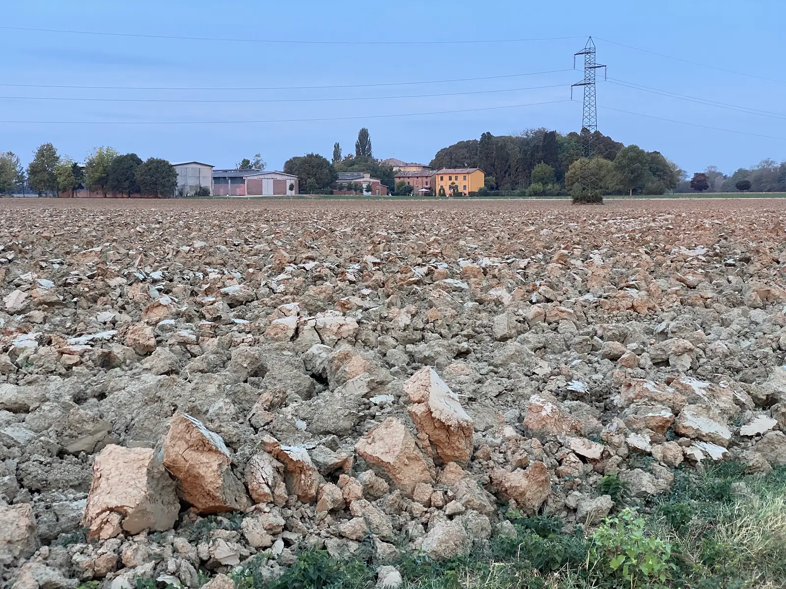 Large clods of freshly ploughed clay soil in the foreground, with farm buildings and an electricity pylon in the distance under a pale evening sky.