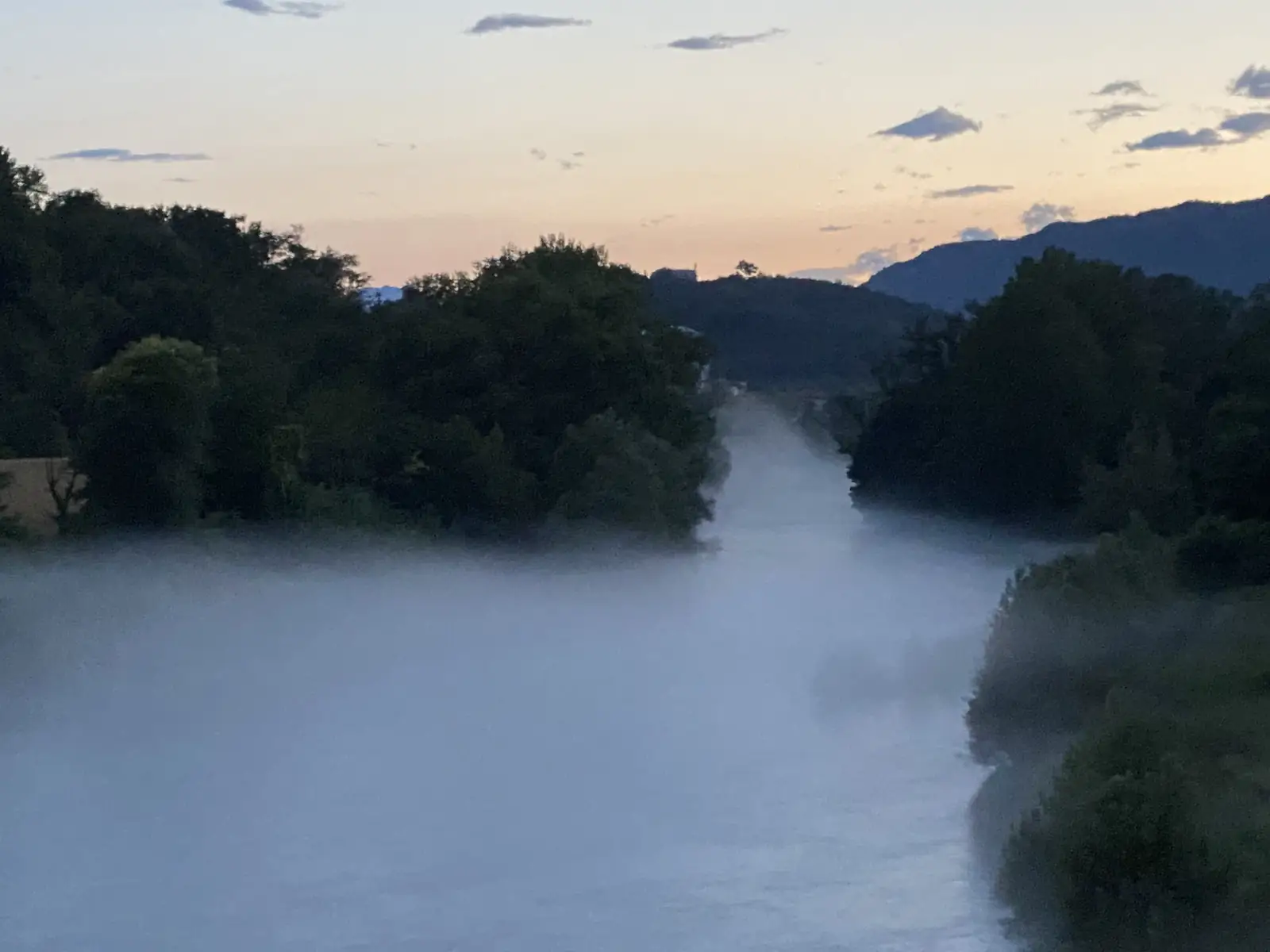 A river valley filled with low mist at dusk, wooded banks on either side and mountains silhouetted against a fading orange sky.