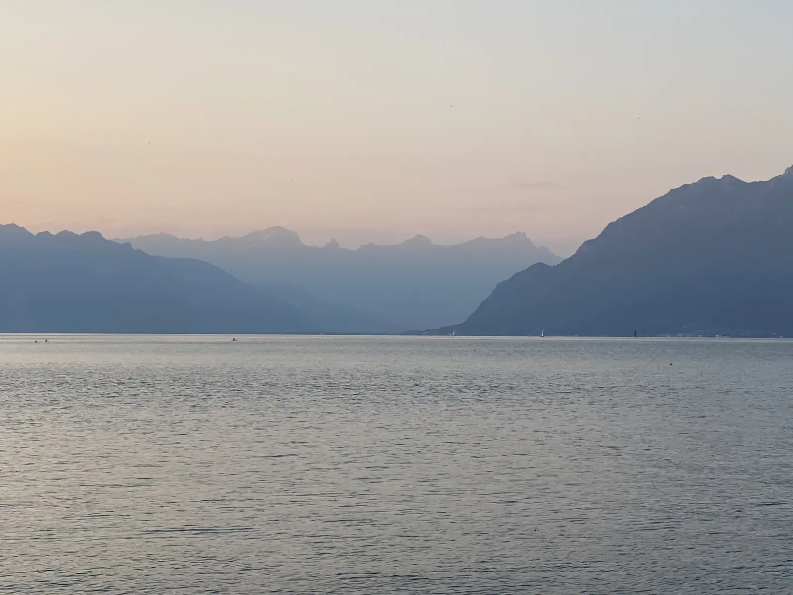 Dusk over Lake Geneva, sailboats silhouetted on the calm water beneath jagged Alpine peaks fading into haze.