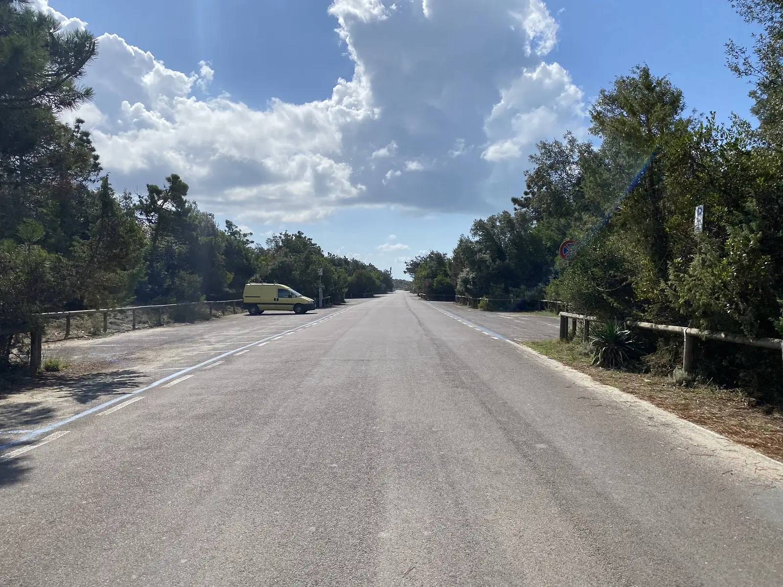 An empty pine-lined coastal road with a single parked van and a no-entry sign, leading towards the shore.
