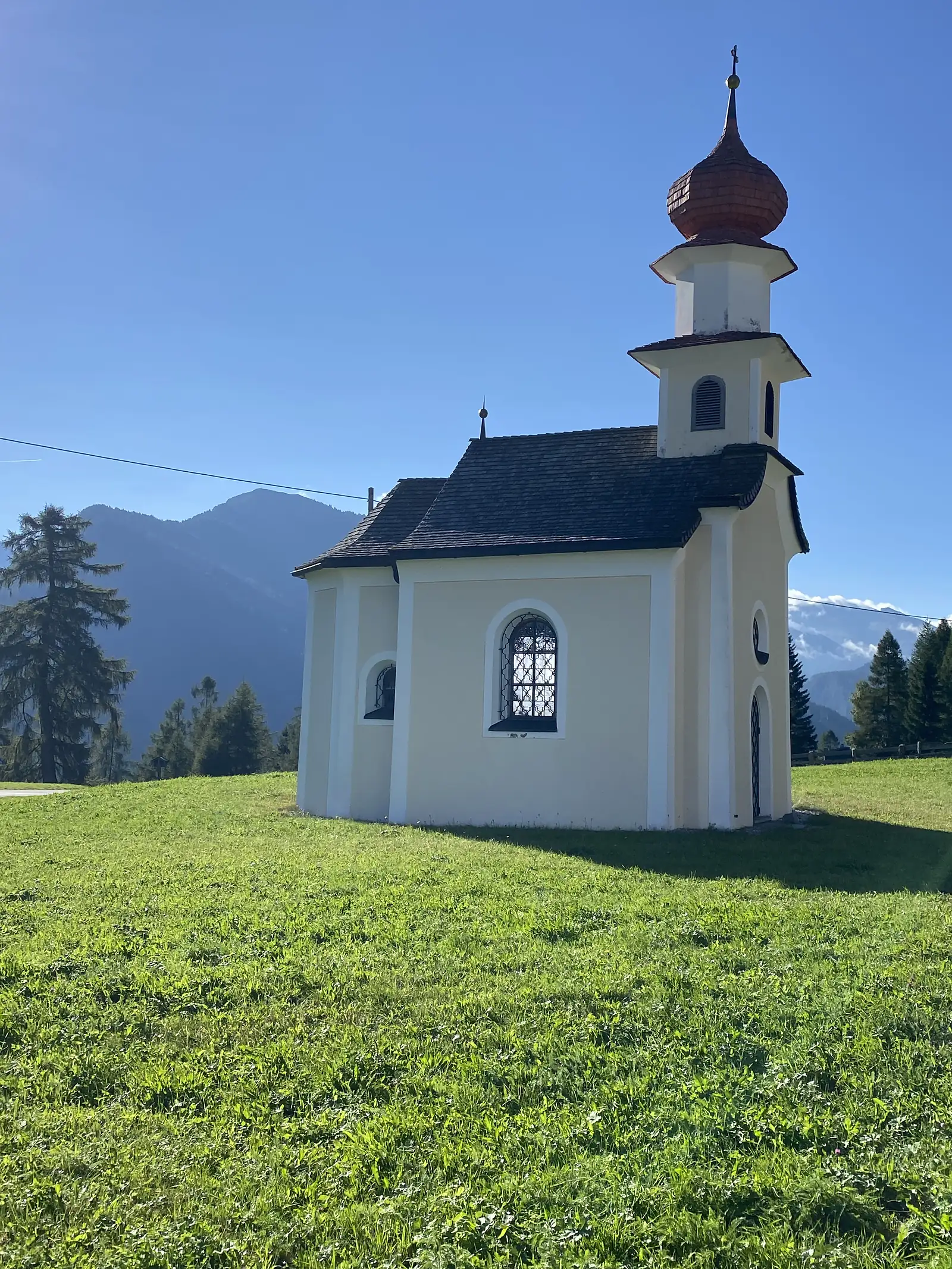 A small white chapel with a copper onion dome and shingled roof stands alone in a grass meadow, with hazy mountains behind.
