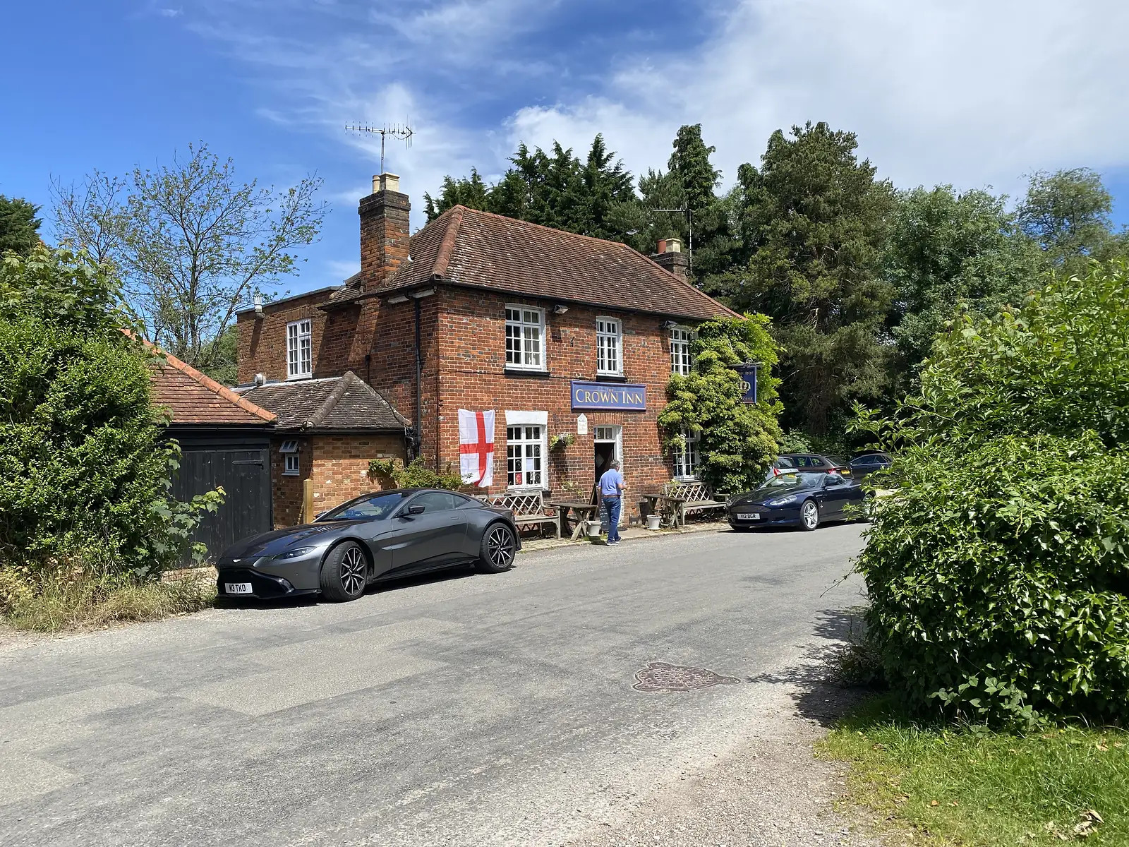 A red-brick tiled country pub lettered "CROWN INN" with a St George's flag on its front and sports cars parked outside.
