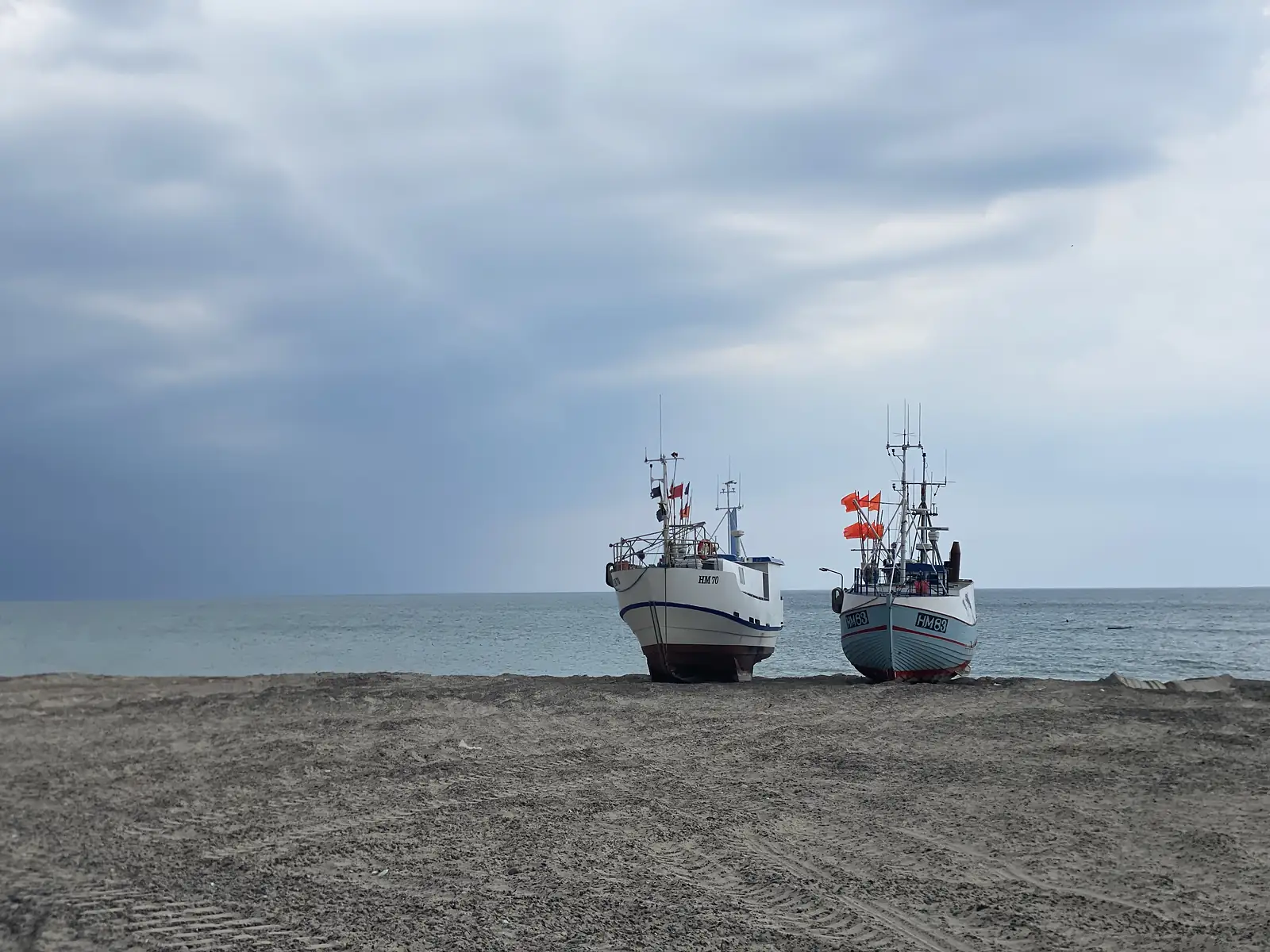 Two beach-launched fishing boats lettered "HM70" and "HM83" standing at the water's edge with orange marker flags, under a rain squall.