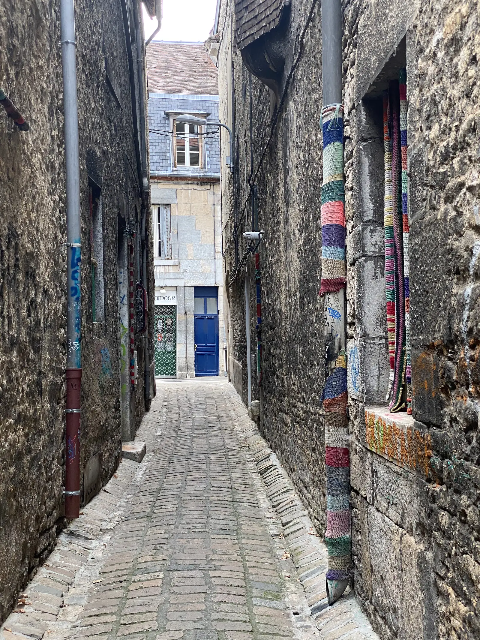 A narrow cobbled alley between old stone houses is wrapped with colourful knitted yarn bombing on a drainpipe and window ledge, leading to a blue door.