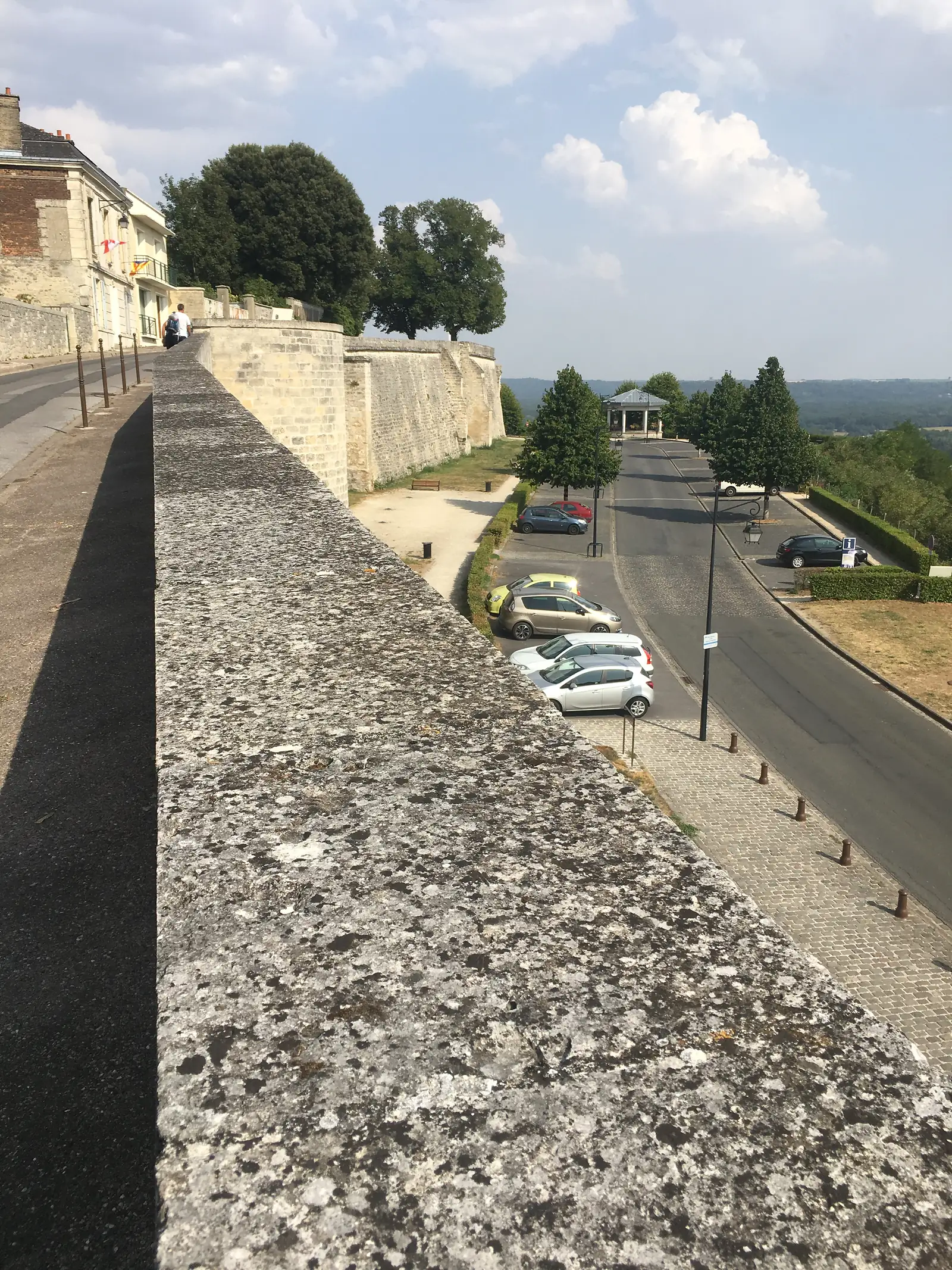 A thick stone rampart wall follows a curving road lined with parked cars and clipped hedges, with the town spreading out below in the haze.