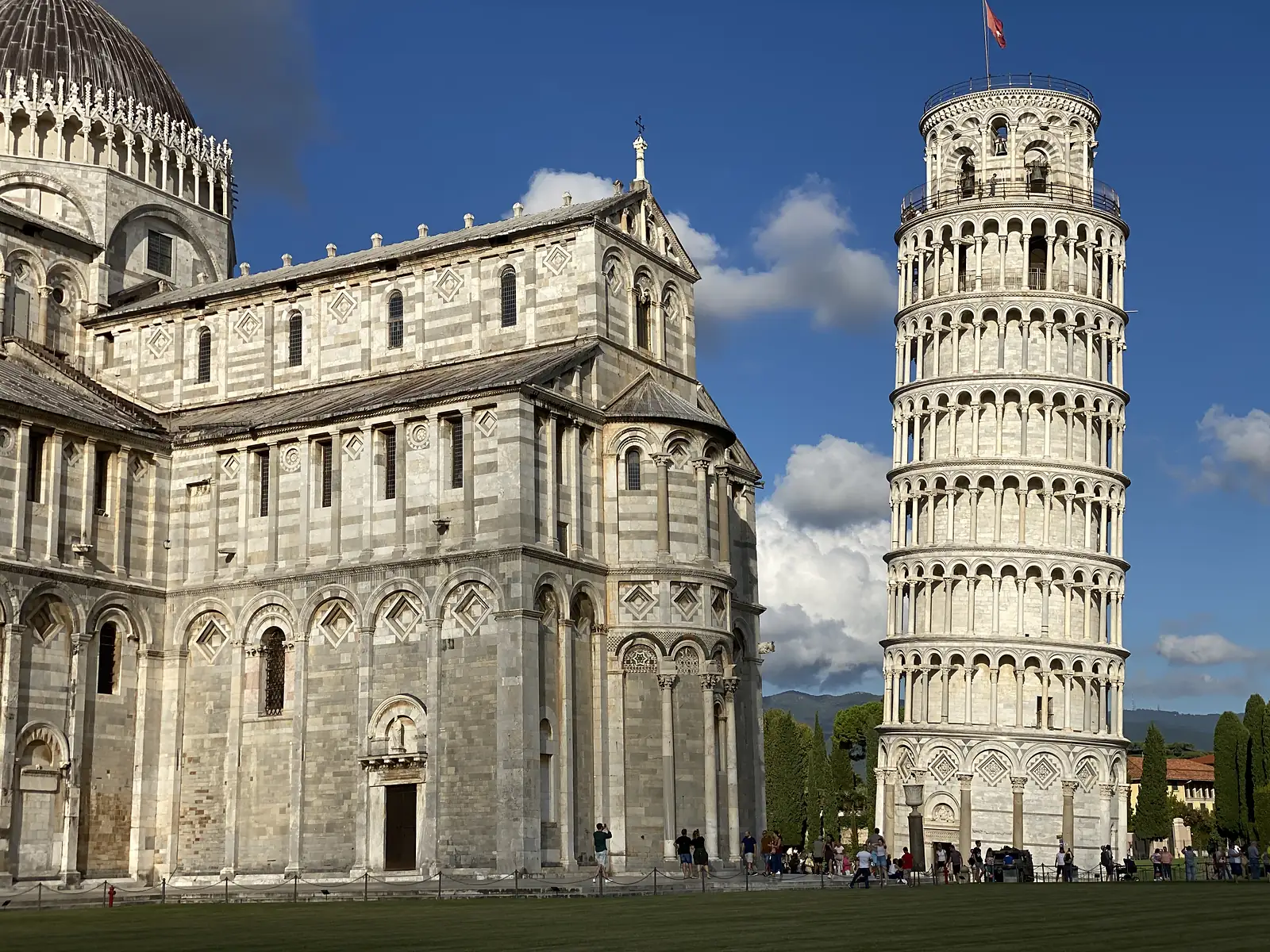 A closer view of Pisa Cathedral's apse and the adjoining Leaning Tower, its marble arcades leaning noticeably above a hedge-lined path.