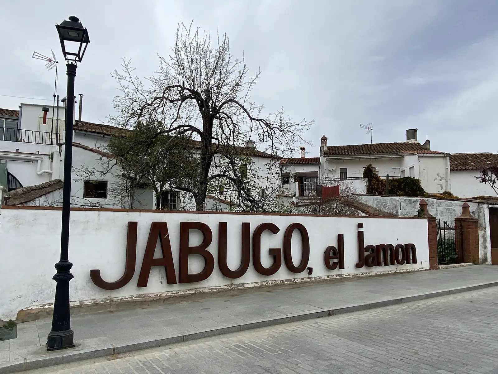 Large rust-coloured metal letters spelling "JABUGO, el Jamon" fixed to a whitewashed wall below a row of village houses.