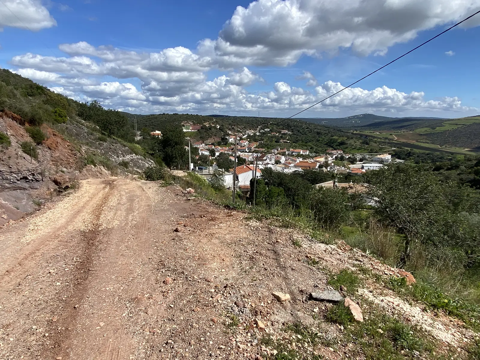 Rough gravel track descending a hillside towards a white village of terracotta roofs spread along the valley floor, with cultivated fields and hills beyond.
