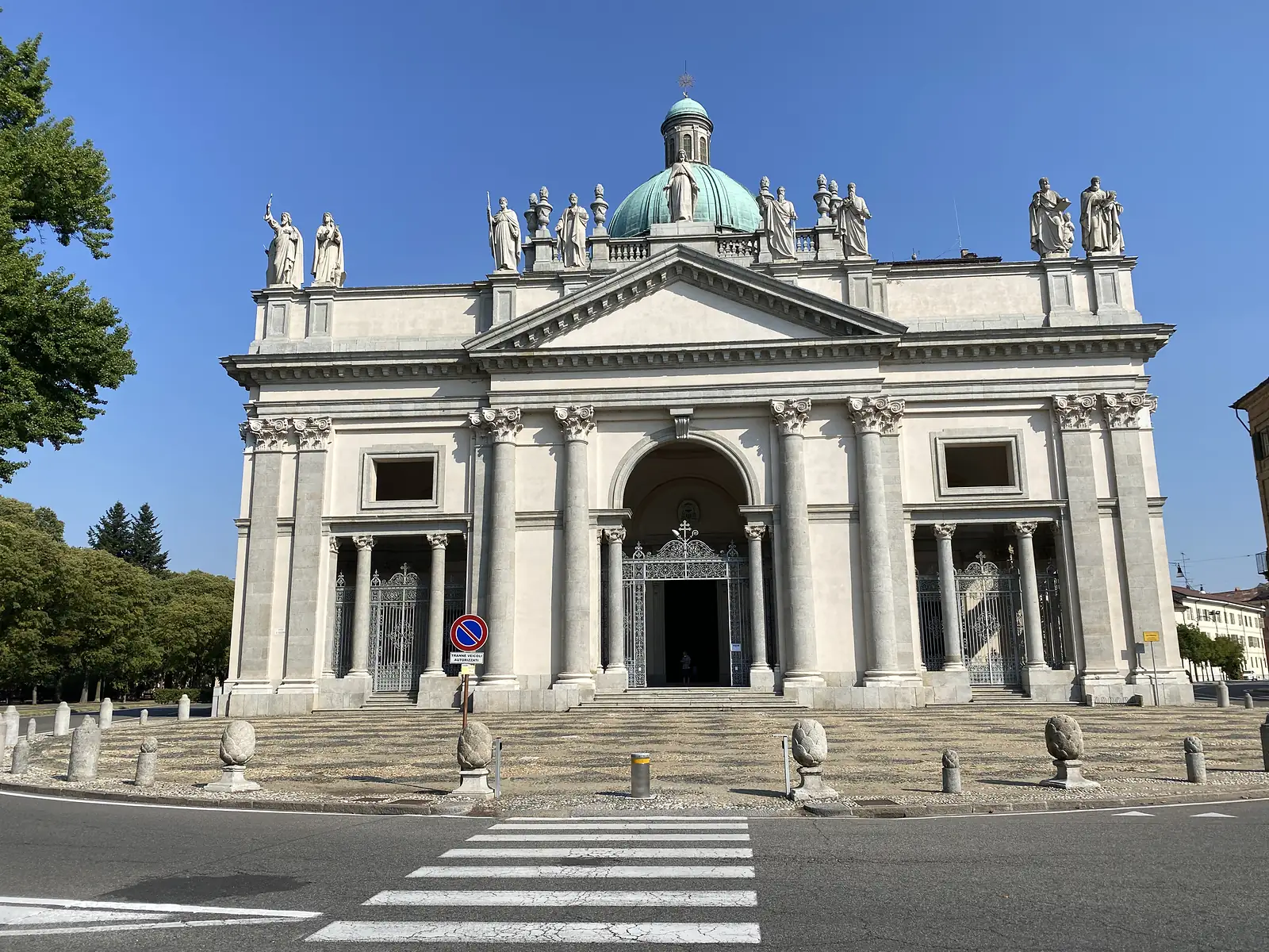 The pale stone facade of Vercelli Cathedral, its pedimented Corinthian portico topped with a row of statues and a green-domed tower, fronted by an empty pedestrian crossing.