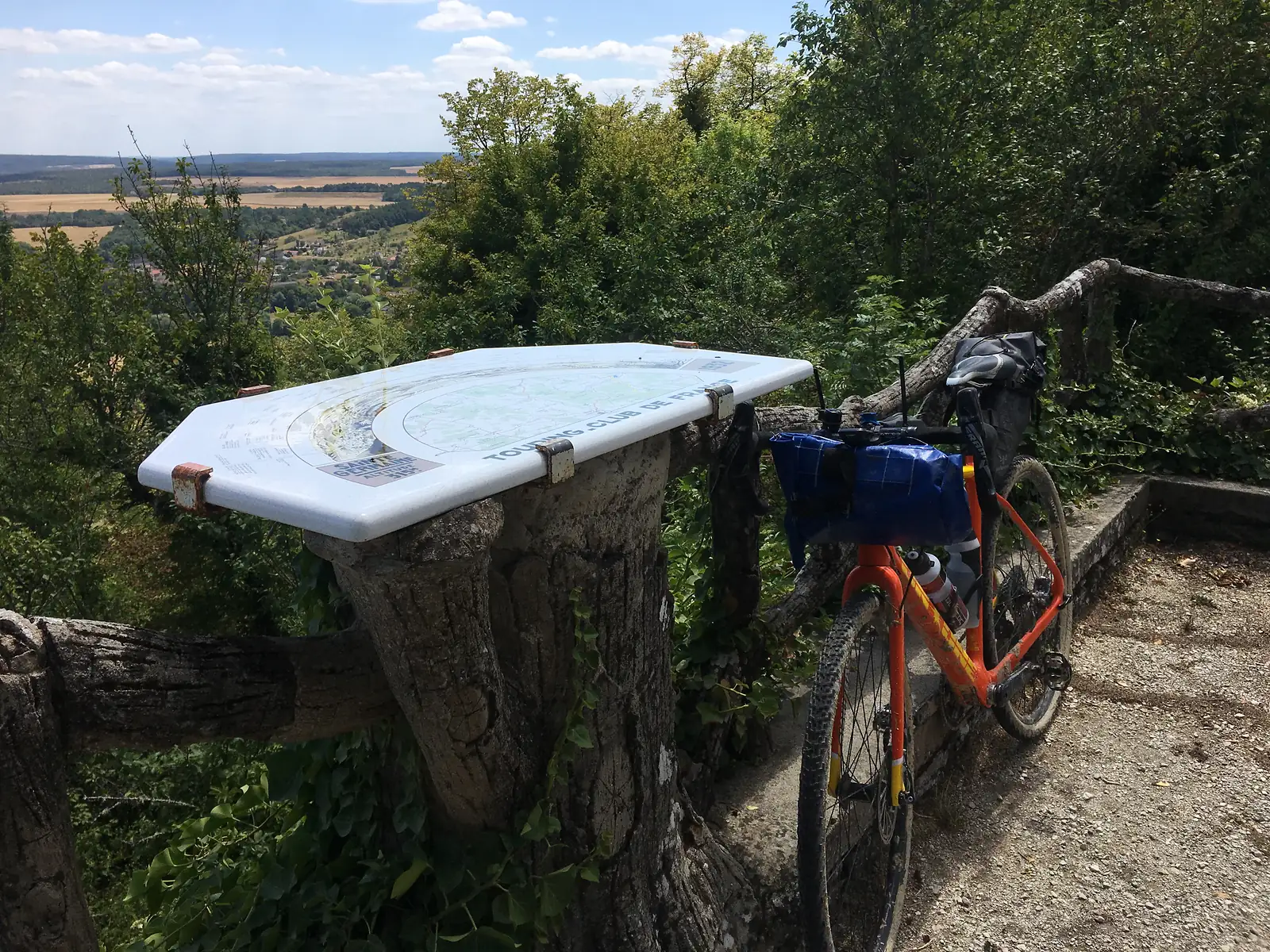 A weathered Touring Club de France orientation table, mounted on a stone plinth, overlooks wooded hills with a loaded bike leaning against the railings beside it.