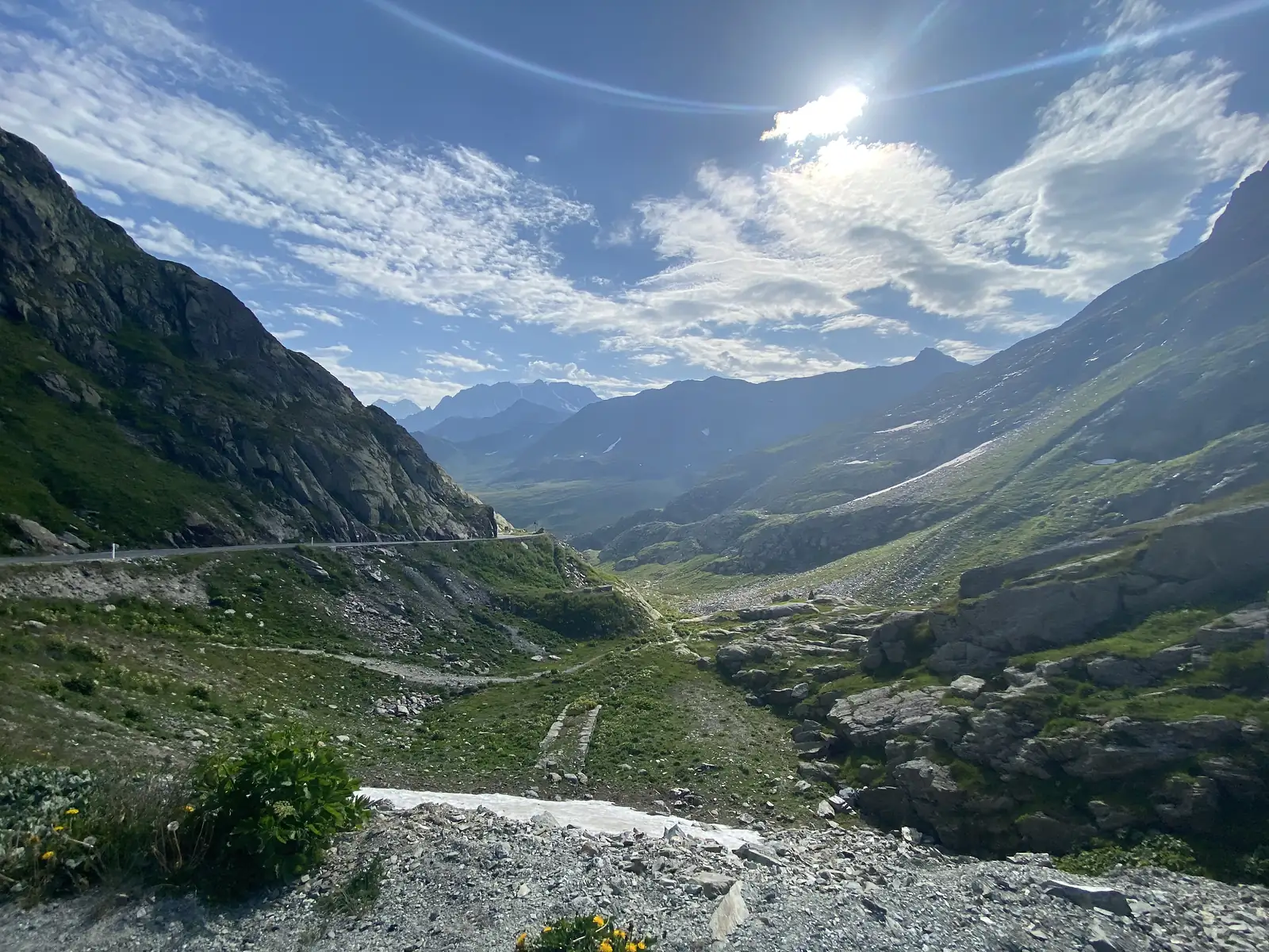 Sunlight streams over a mountain ridge above a rugged, snow-patched valley, a stone-paved path leading down towards the road.