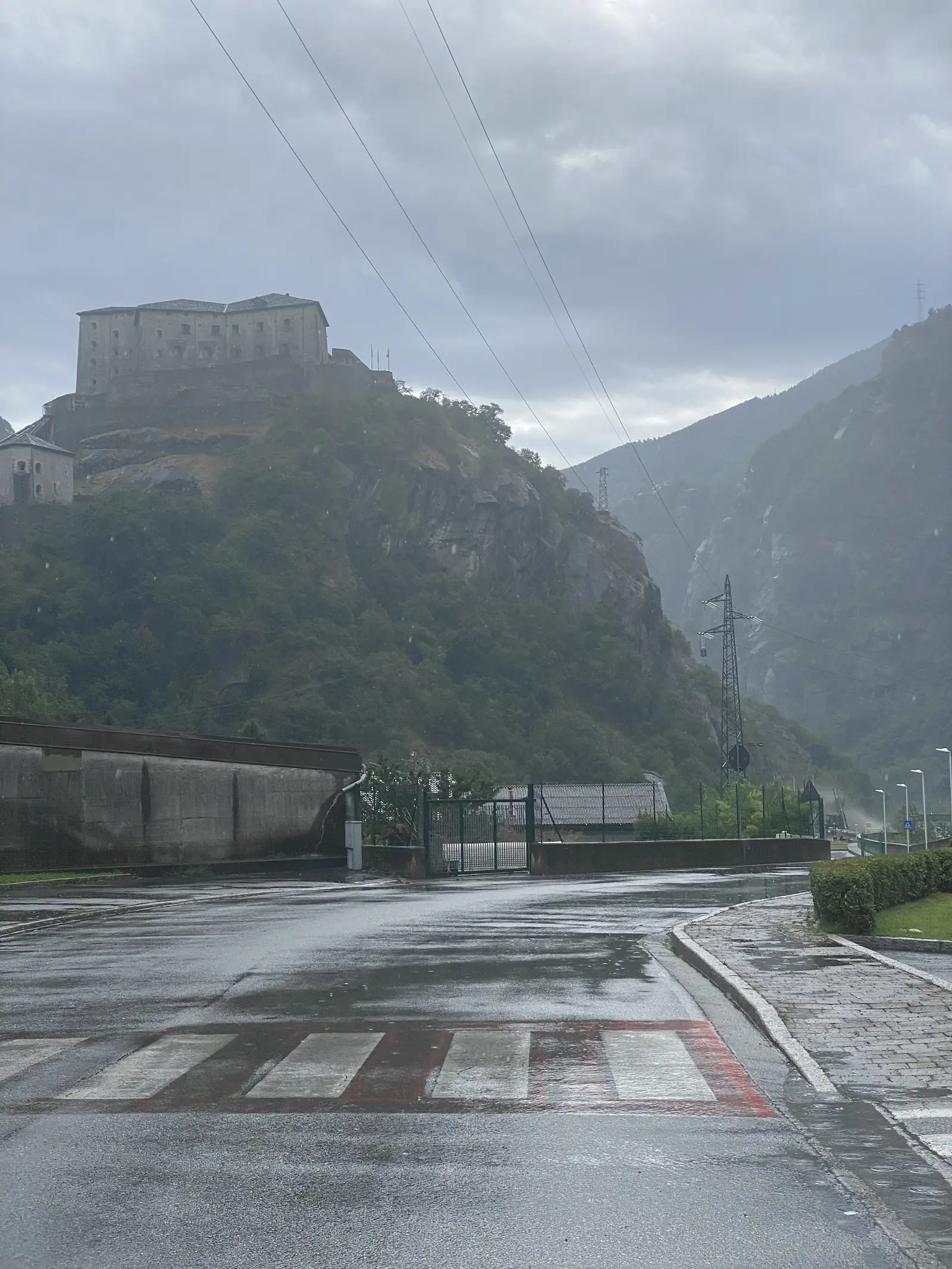 The multi-tiered Fort of Bard crowning a rocky outcrop above a rain-wet road junction with a zebra crossing, seen from below on a grey day.