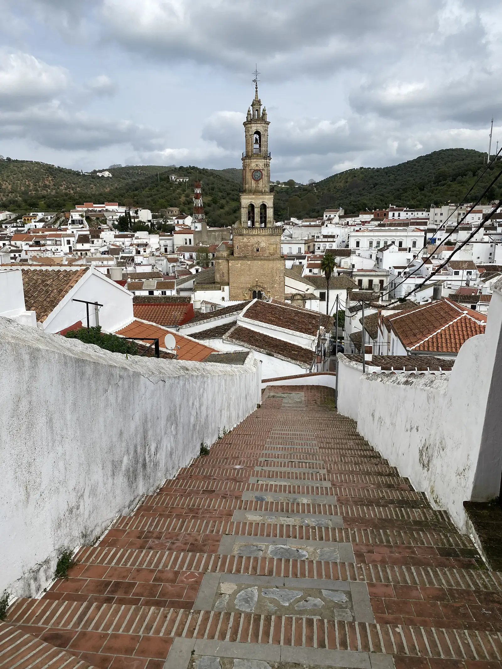 Brick-paved steps descending between whitewashed walls towards a town of white houses and red roofs, dominated by a tall stone bell tower with a clock face.