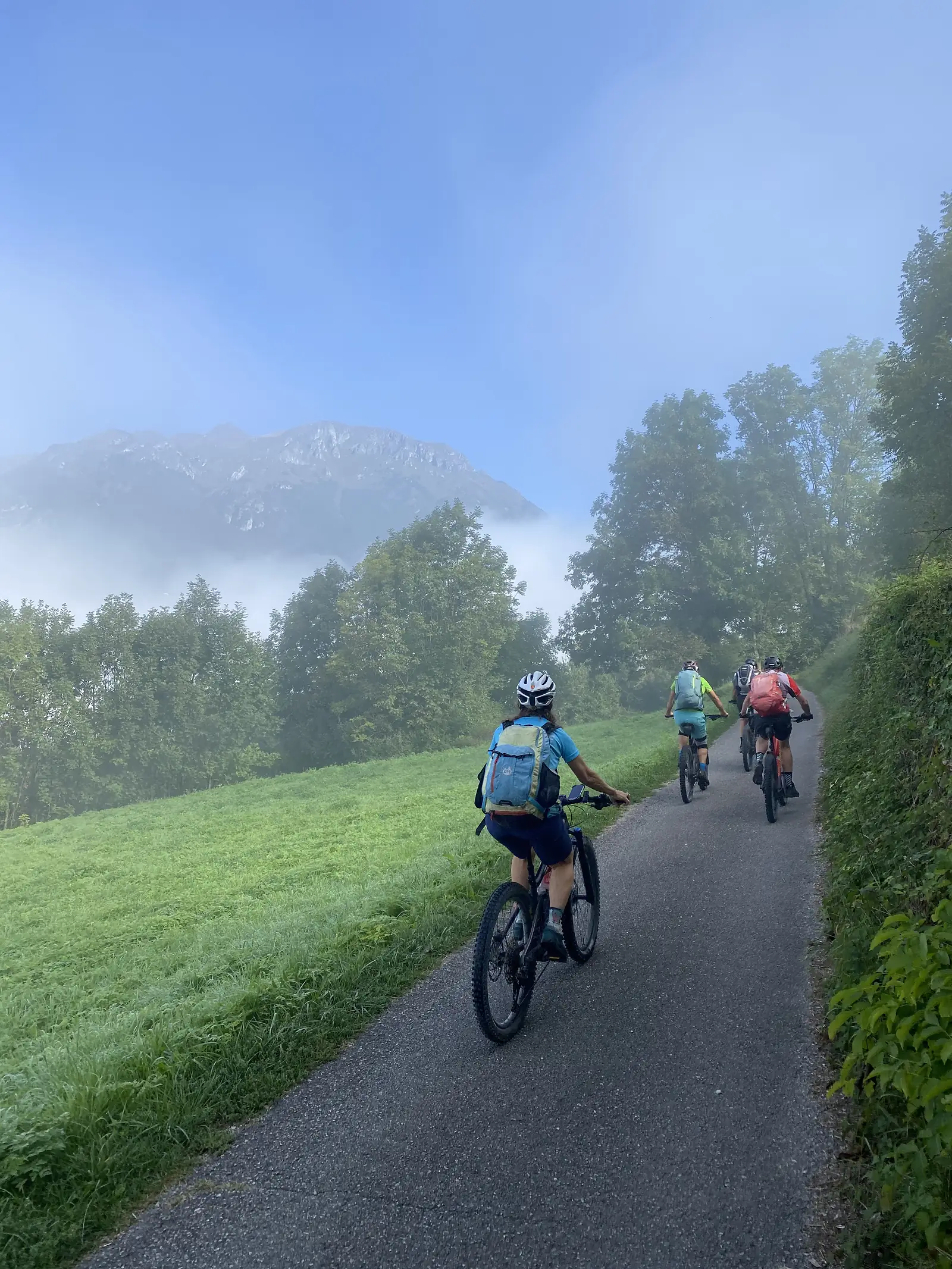 Three riders on mountain bikes with rucksacks climb a narrow lane, with mist lying along the foot of a rocky ridge ahead.