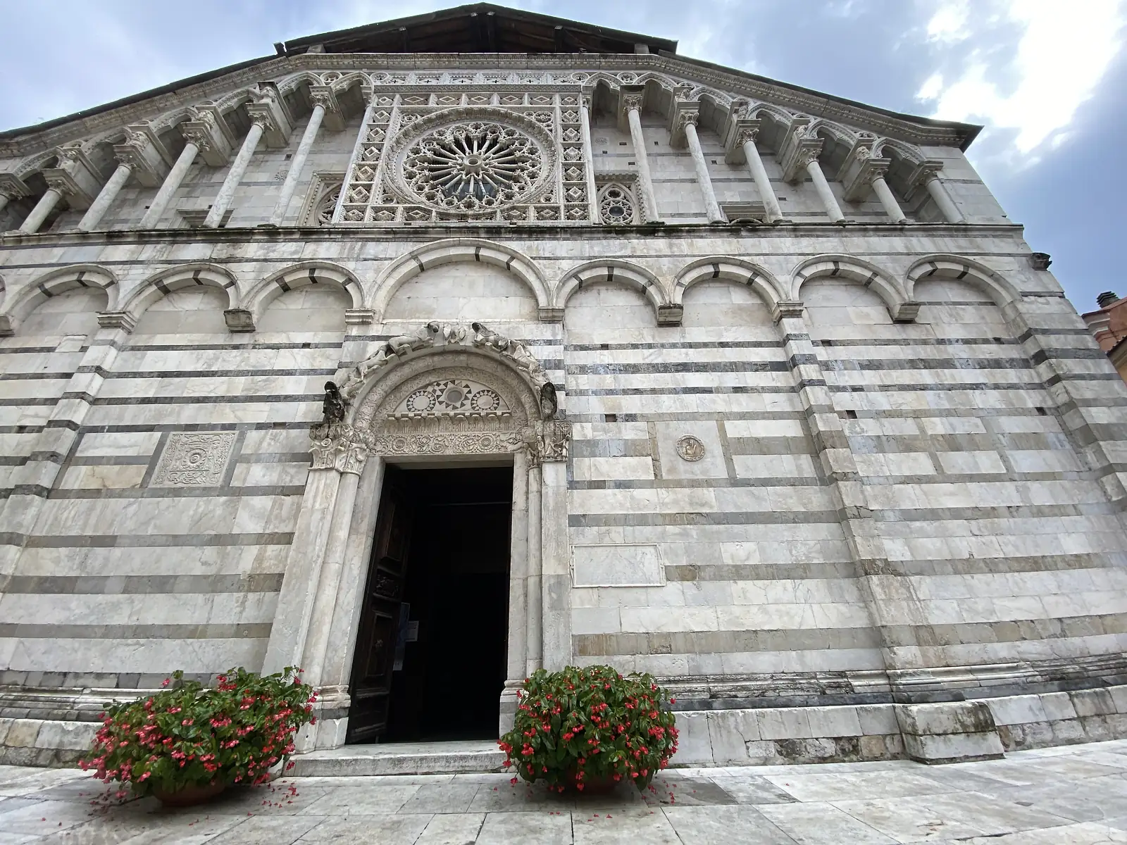 A striped grey-and-white marble church façade with a large carved rose window and an ornate marble portal, its steps flanked by pots of red flowers.