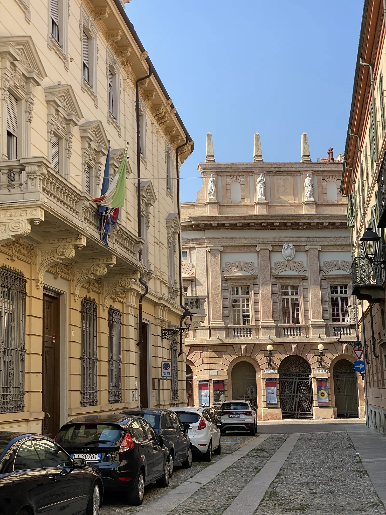 An ornate cream stucco townhouse in Ivrea, its wrought-iron balconies overhung by a fluttering Italian flag, with cars parked along the pavement below.