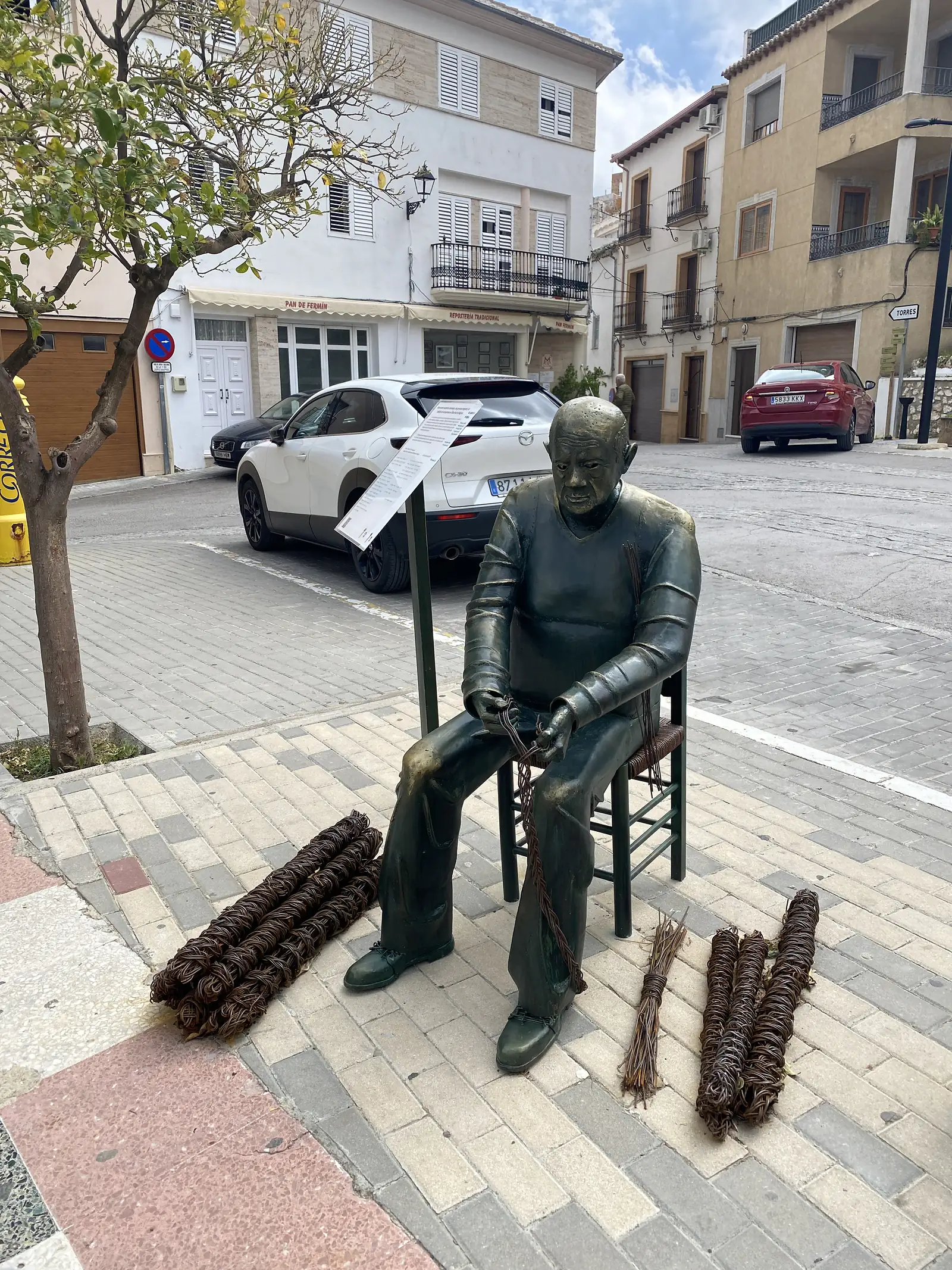 A bronze statue of a seated man weaving a rush chair seat on a village pavement, bundles of cane laid at his feet.