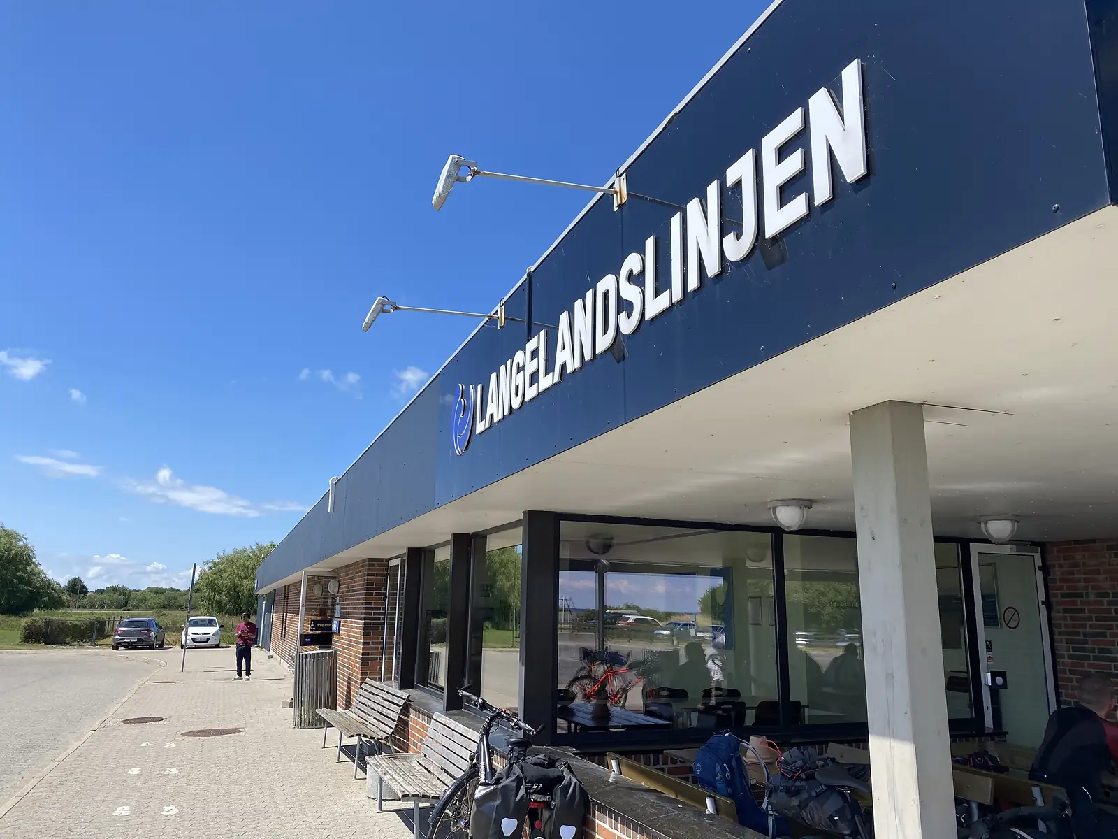 A ferry terminal lettered "LANGELANDSLINJEN" under a blue sky, with loaded touring bikes and luggage on benches outside.