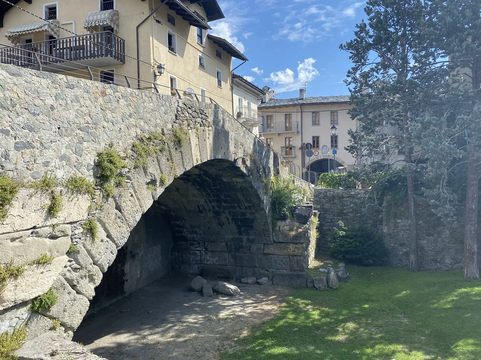 An old single-arch stone bridge spans a dry gully beside a cluster of pastel townhouses, pine trees and a road sign visible on the far bank.
