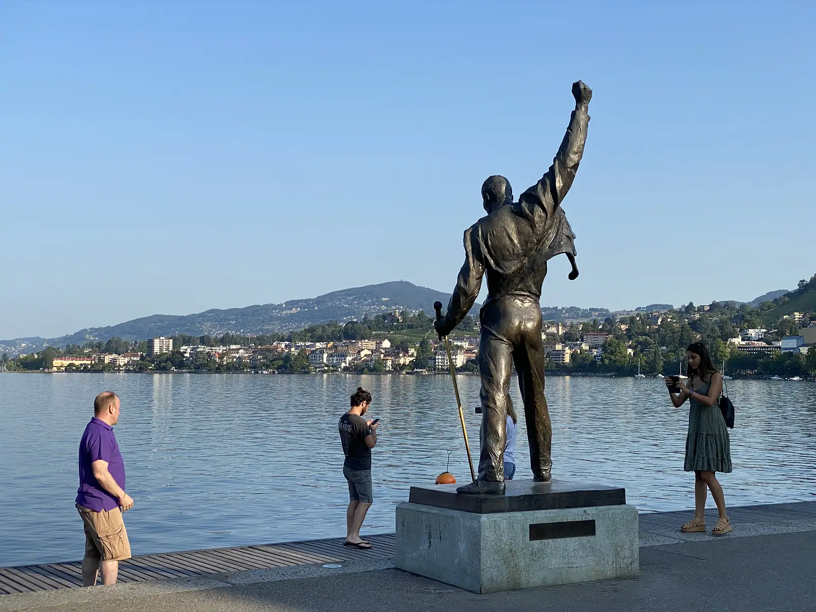 Three onlookers photograph a bronze statue of a performer with one fist raised skyward on a lakeside promenade, the town and hills across the water behind.