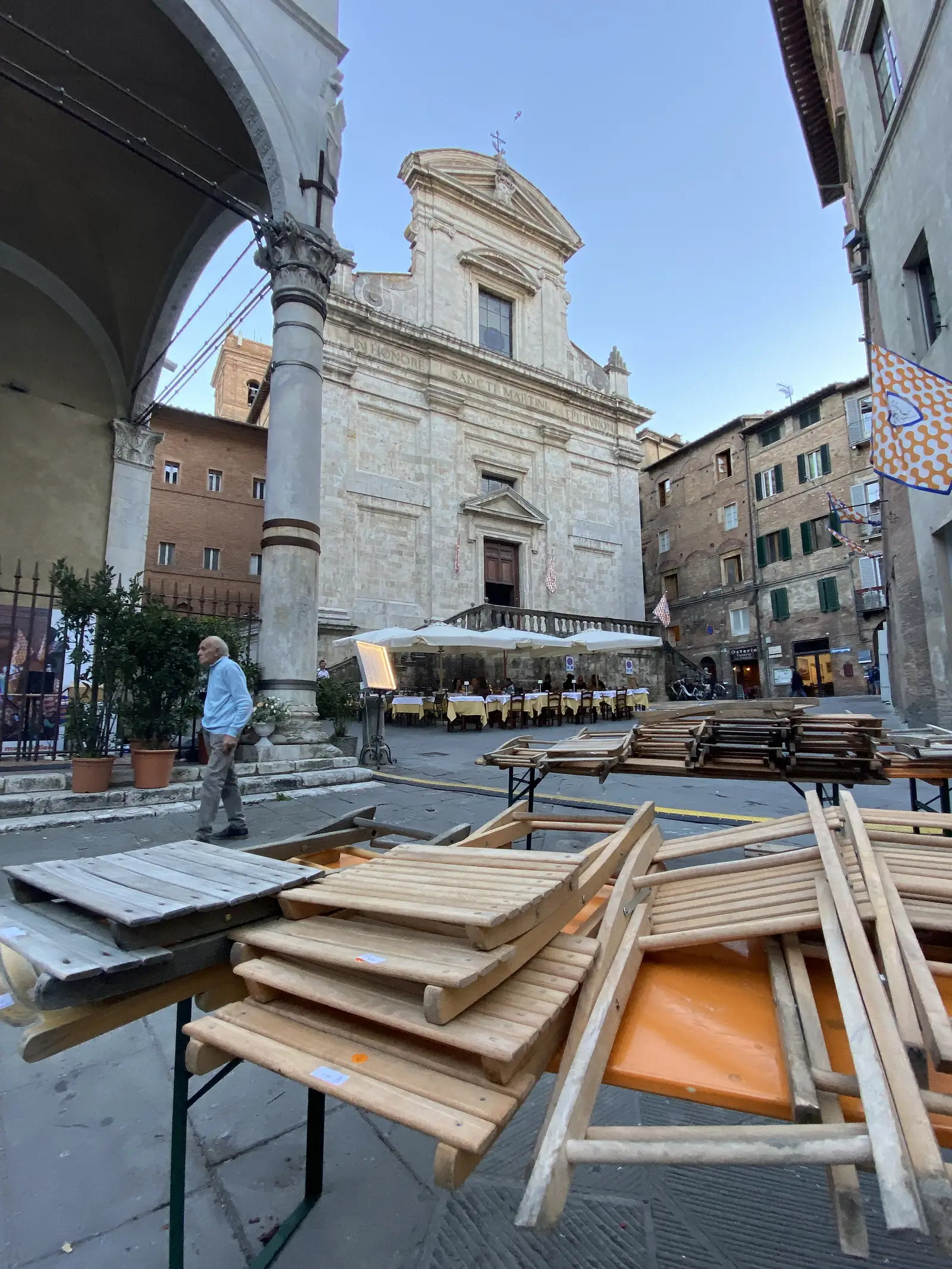 A baroque church facade inscribed "IN HONORE SANCTI MARTINI", overlooking a piazza lined with restaurant tables and stacked wooden chairs.