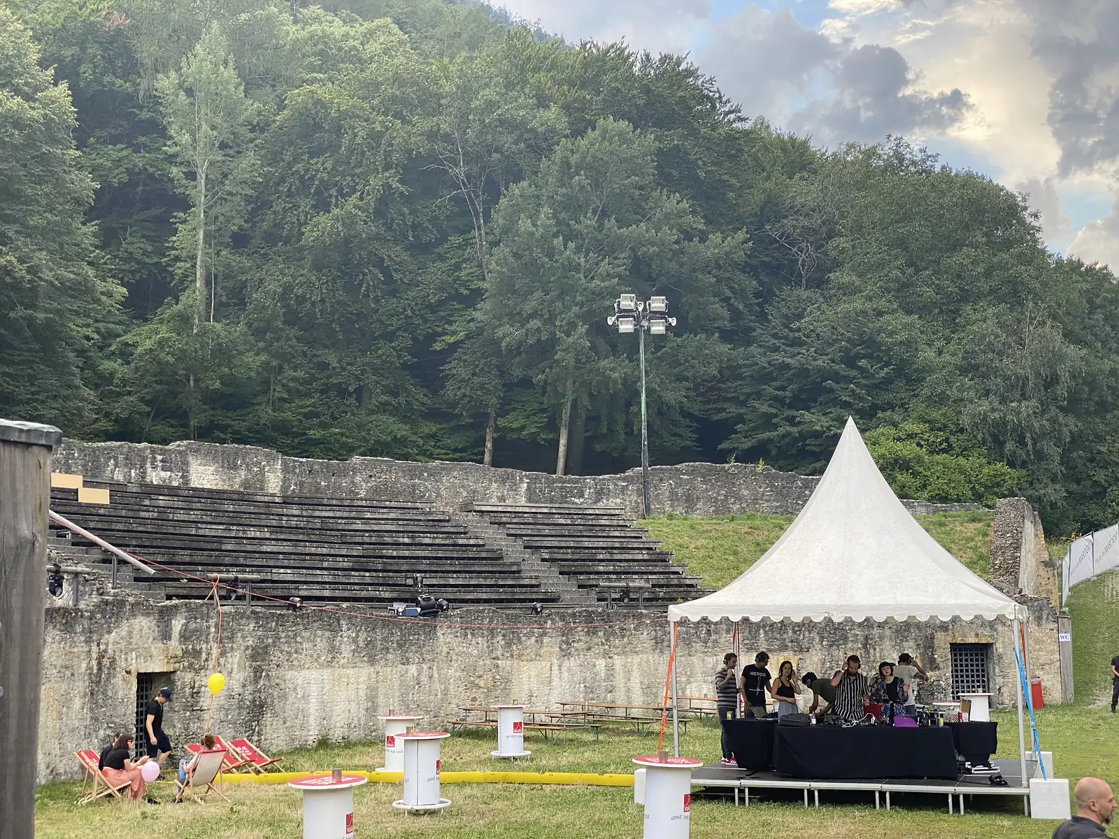 The weathered stone tiers of an ancient open-air amphitheatre host a small outdoor event, with a marquee, DJ setup and deckchairs set against a wooded hillside.