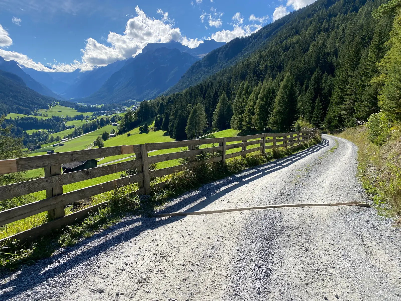 A gravel track runs beside a timber post-and-rail fence above a broad green valley, with a jagged rock ridge closing the head of it.