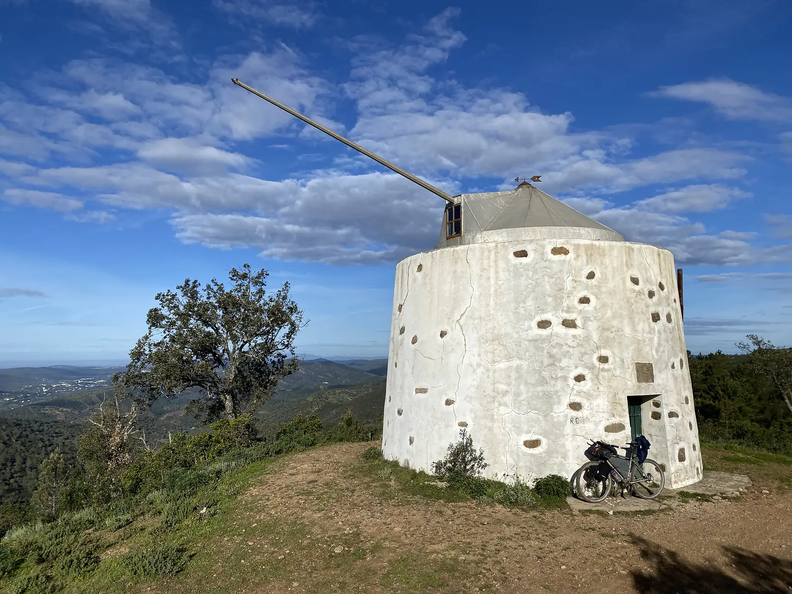 Whitewashed stone windmill tower on a hilltop, one long bare sail spar still in place, a loaded bike leaning against its wall and wooded valleys falling away behind.