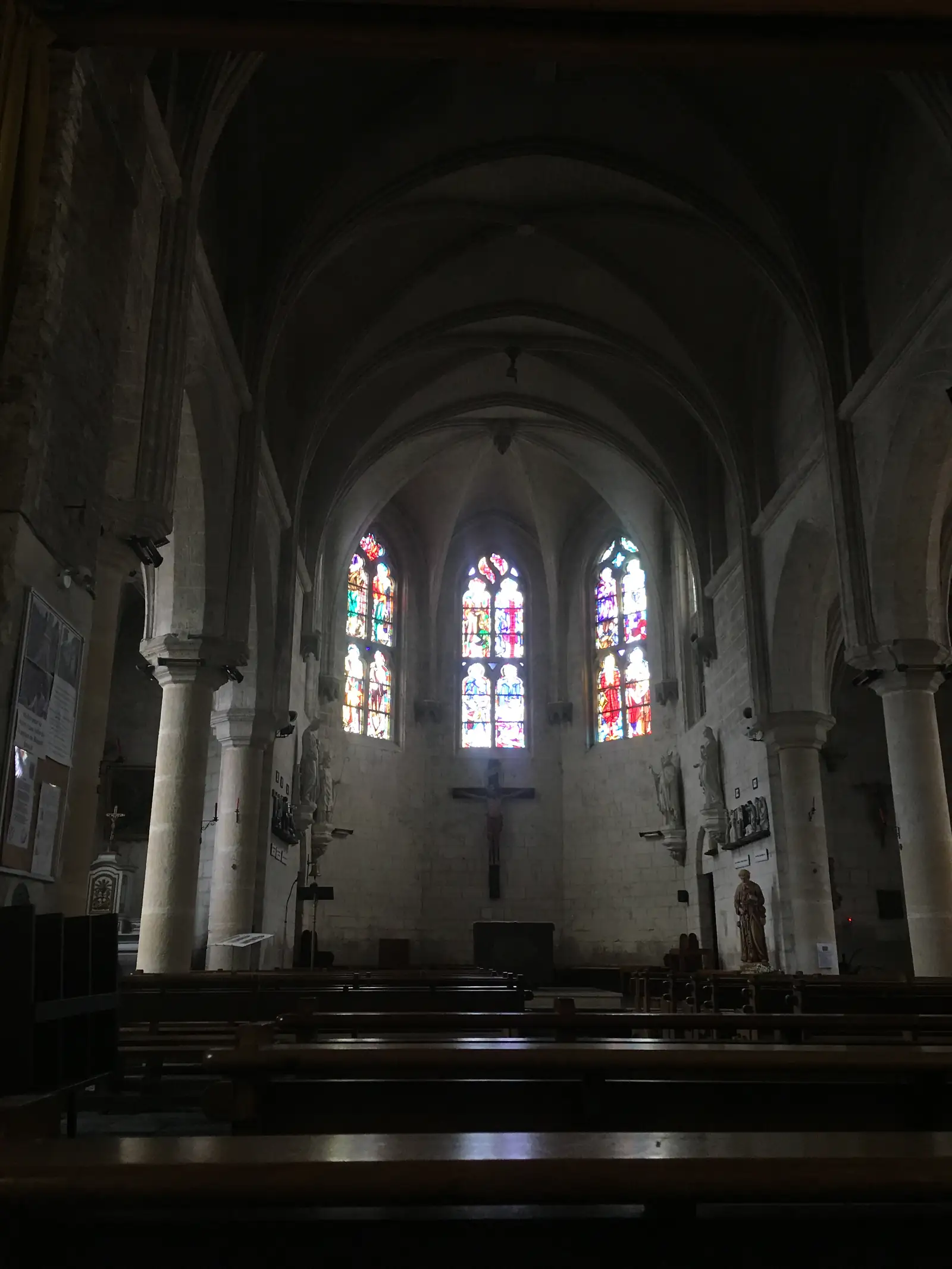 Sunlit stained-glass windows illuminate the nave of a Gothic church, with a hanging crucifix and stone columns rising into vaulted arches.