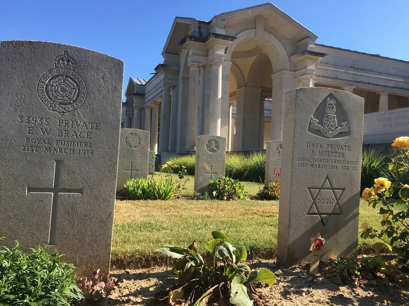 Rows of white headstones in the Faubourg-d'Amiens Cemetery leading to the colonnaded Arras Memorial, poplar trees lining the boundary beyond.