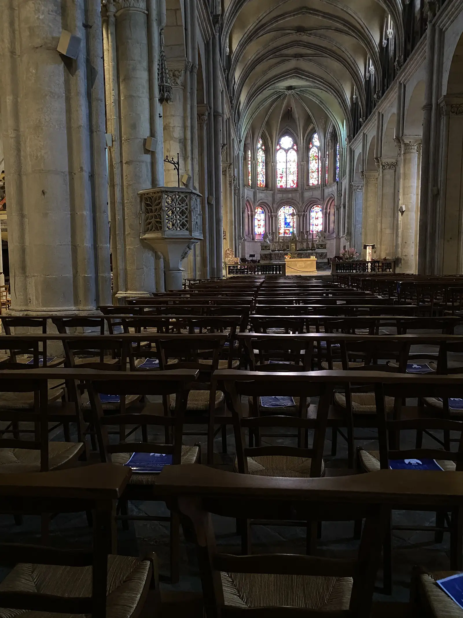 The vaulted stone nave of a cathedral leads past rows of wooden chairs to an apse lit by tall stained-glass windows.