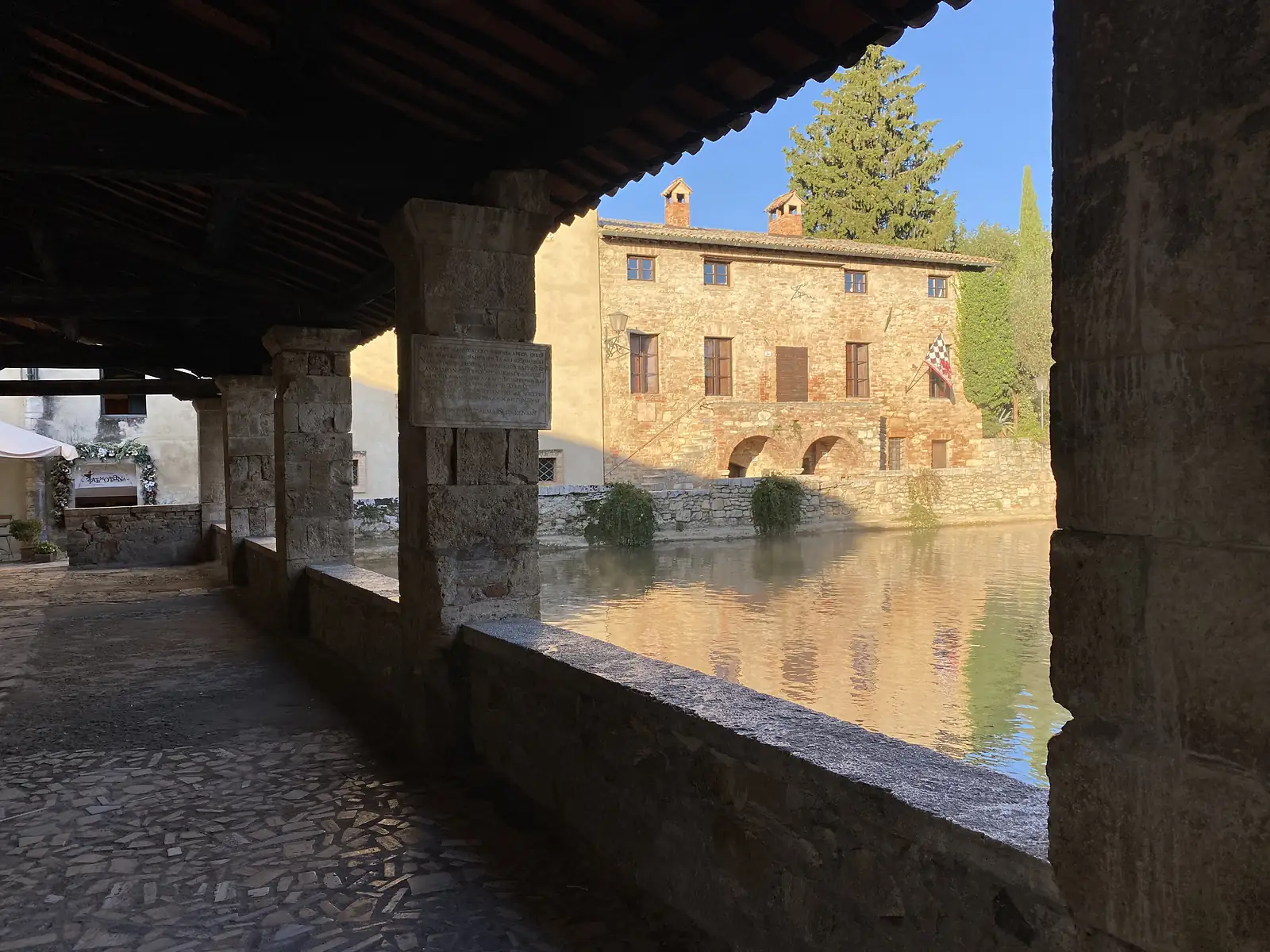 A steaming rectangular thermal pool filling a stone piazza, bordered by a colonnaded walkway and a stone farmhouse with twin chimneys.