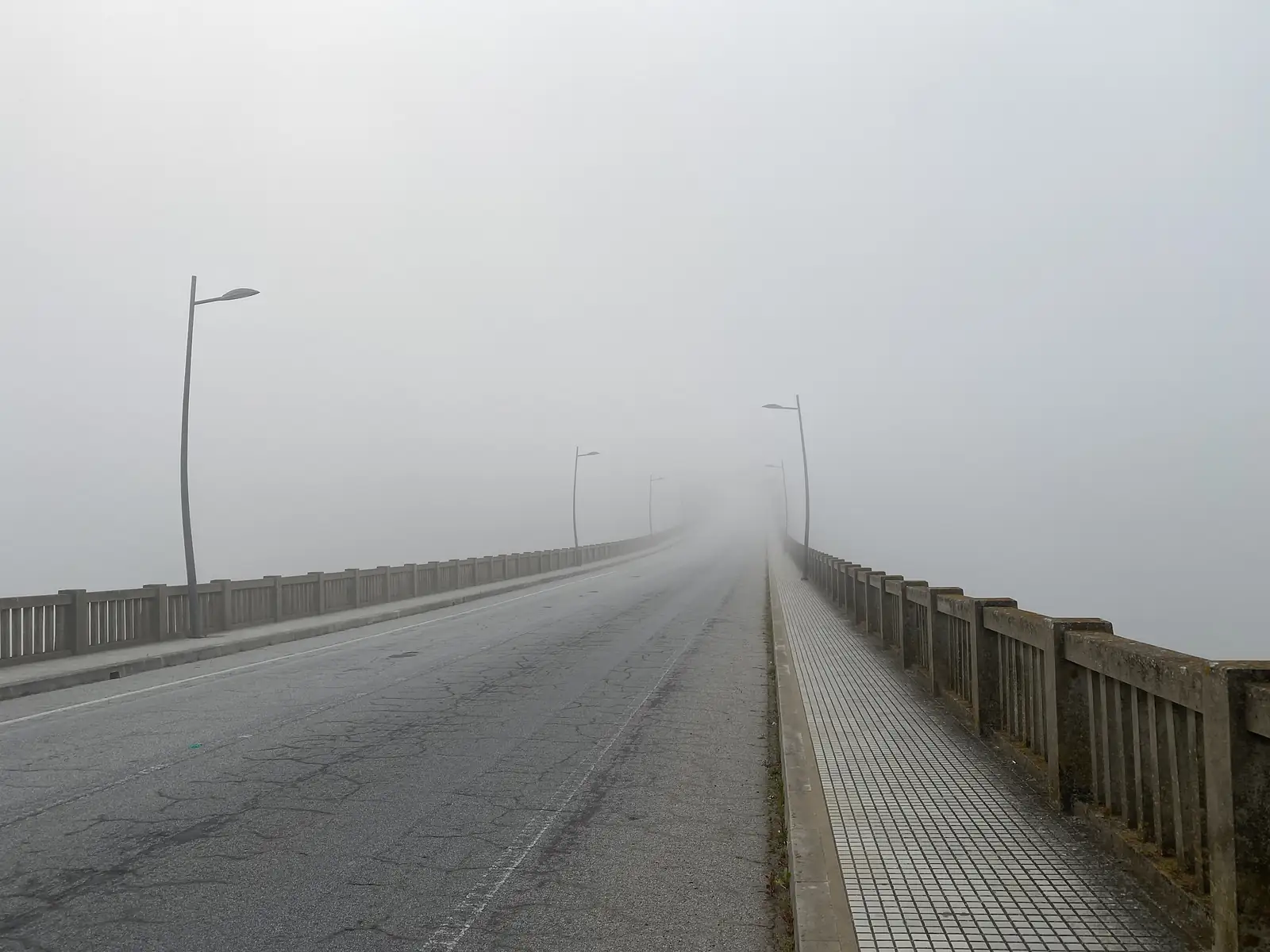 An empty road bridge with a concrete balustrade and a row of street lamps vanishing into thick fog.