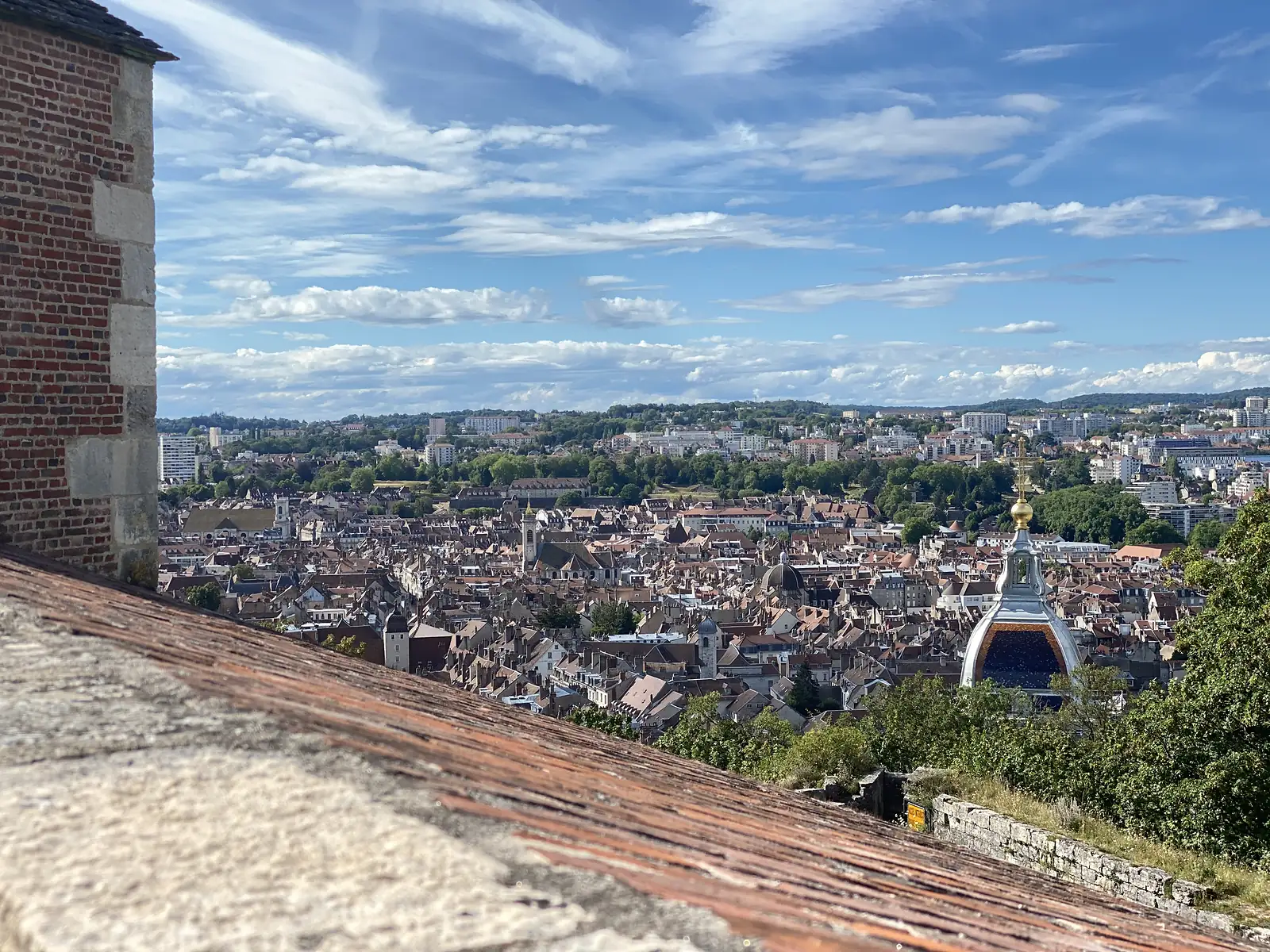 Terracotta rooftops and a domed church spire spread across Besançon, seen from a high vantage point beside a brick and stone wall.