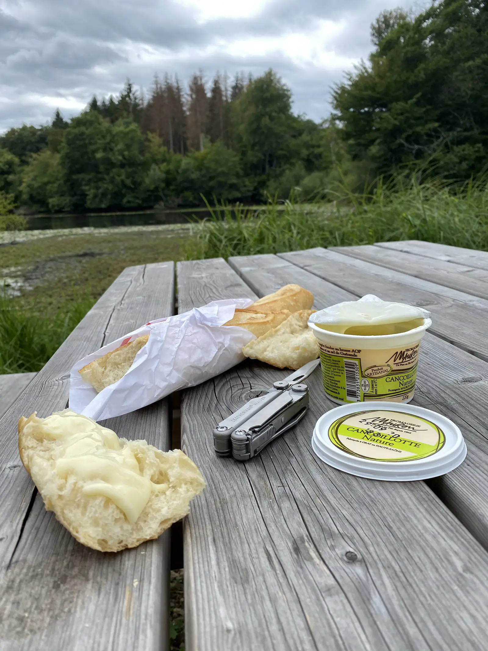 A picnic of torn baguette, butter and a tub of Cancoillotte cheese is laid out with a multitool on a wooden table beside a reed-fringed lake.
